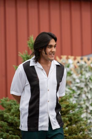 A person in a white and black bowling-style shirt smiles while standing against a red wall outdoors.