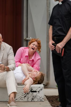 Performers act out an emotional scene on steps while someone in clerical attire looks on.