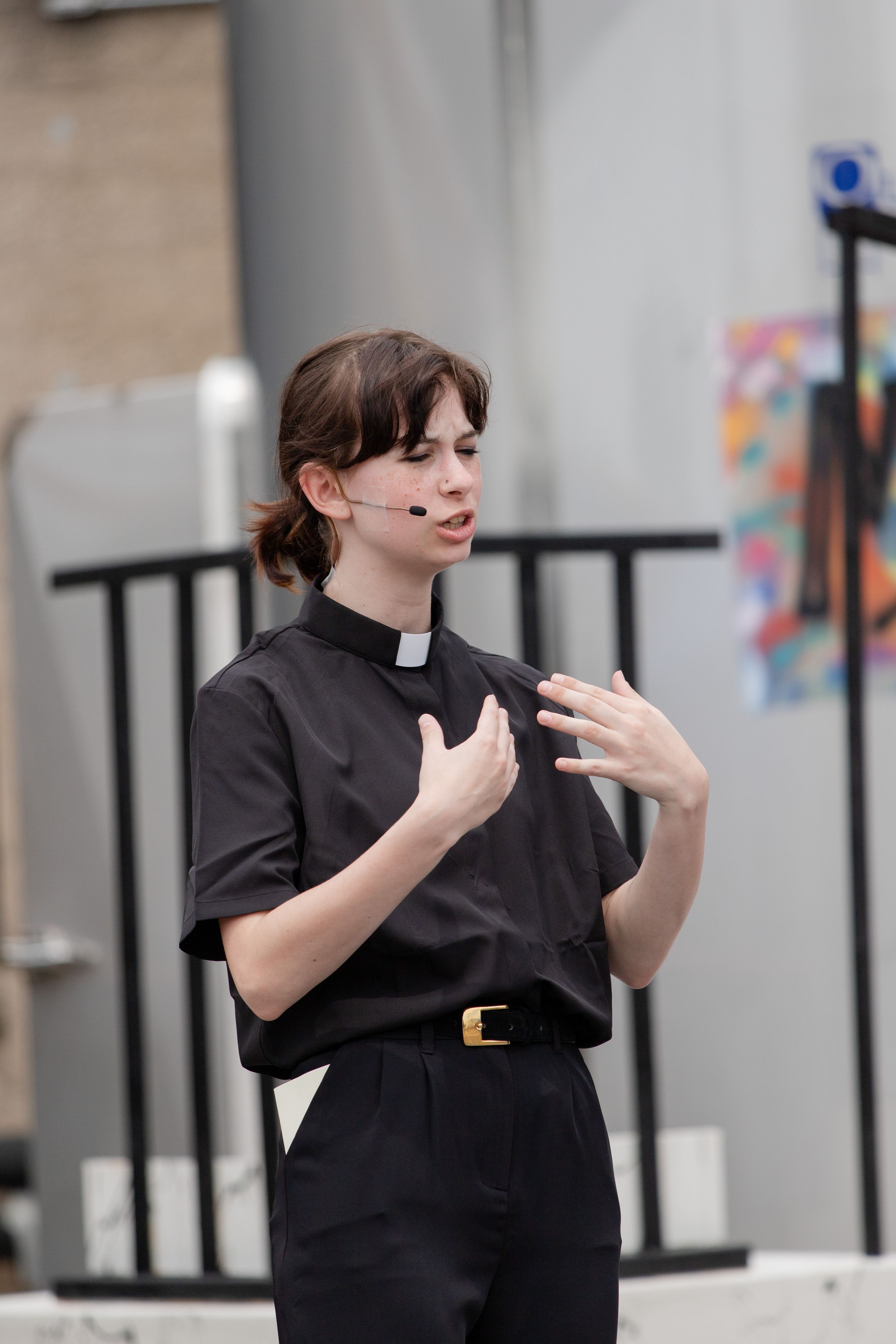 Professional figure in all black attire makes expressive hand gestures while speaking outdoors.