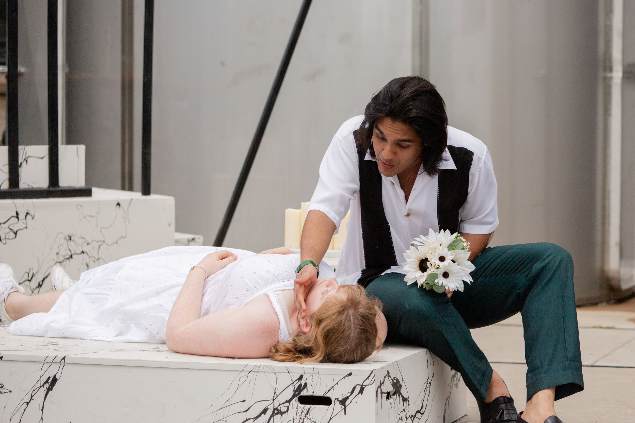 Two people in an artistic performance on stage with white flowers and dramatic lighting.