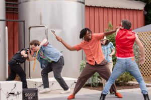 A group of people strike dramatic dance poses in front of a building with red siding and lattice fence.