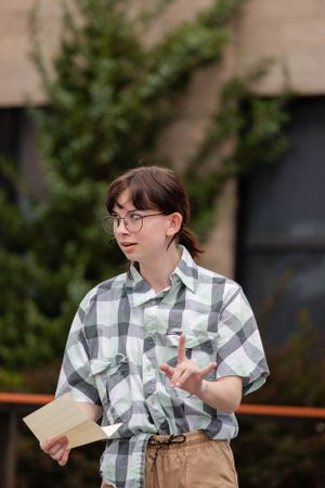 A person in a plaid shirt and glasses holds papers while standing outdoors near greenery.