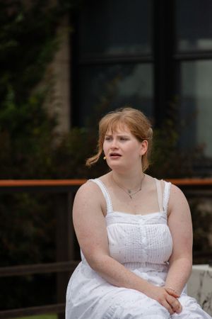 A person in a white sundress sits outdoors on a summer day, looking thoughtfully to the side.