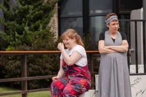 Two people in contrasting moods stand outside a building, one seated in plaid pants while another stands with crossed arms.