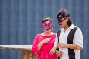 Two people wearing colorful masks share a laugh together against blue wall background.