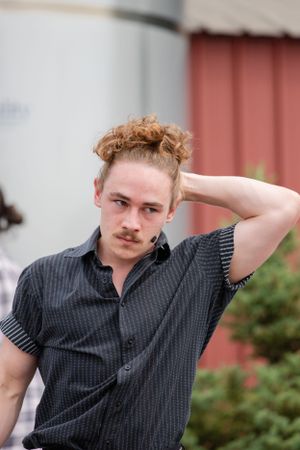 Person in dark striped shirt poses with hand in hair against red and white wall backdrop.