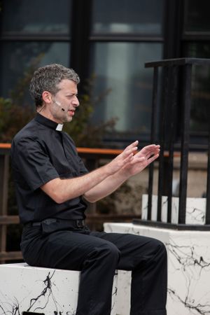 A person in clerical attire gestures while seated on a concrete platform outdoors.