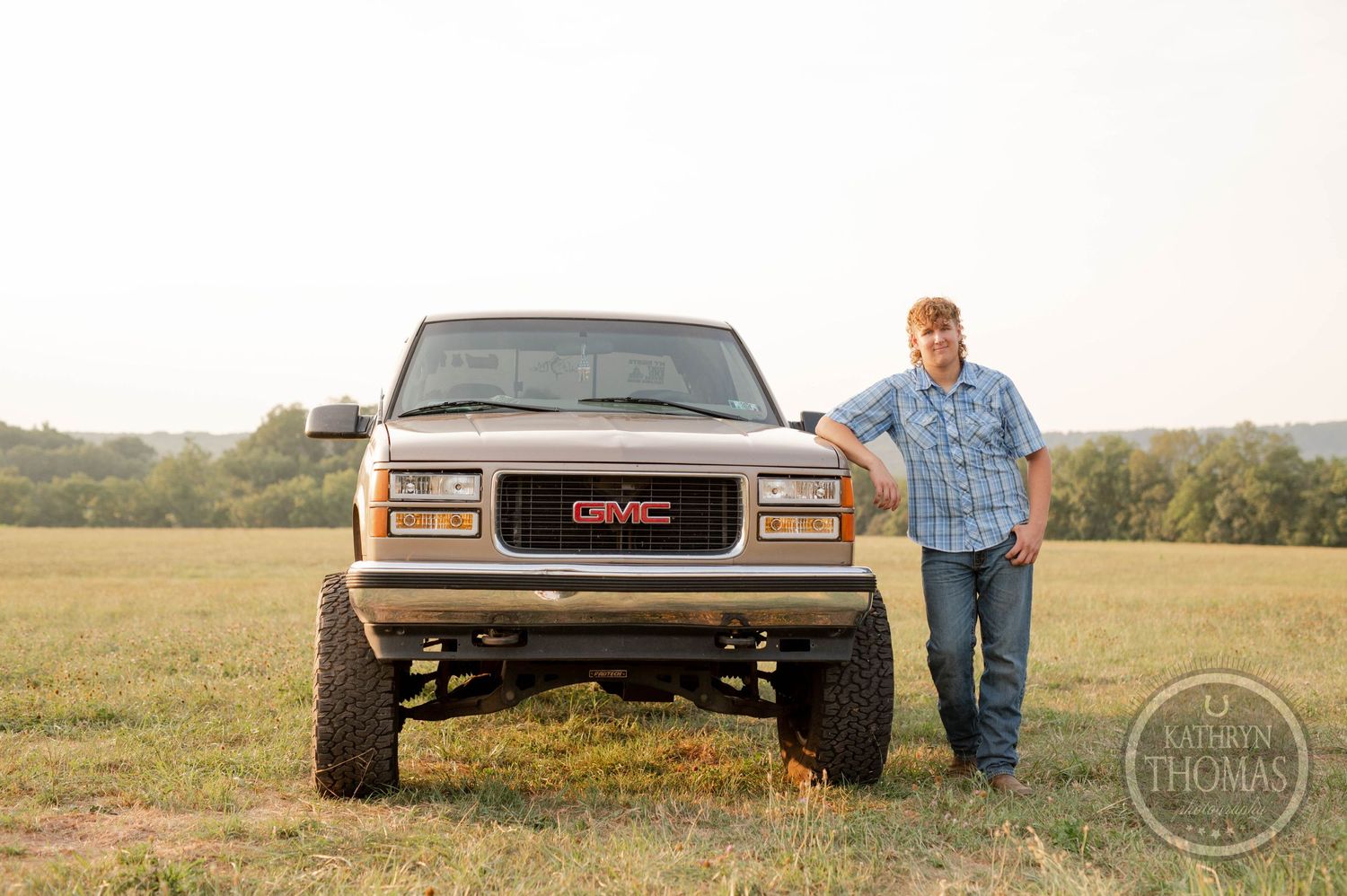 A person posing next to a lifted GMC pickup truck in golden hour lighting.