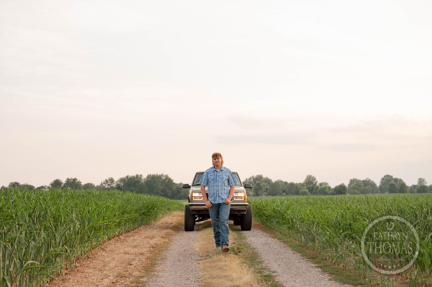Person in blue shirt walking on dirt path between cornfields with truck in background.