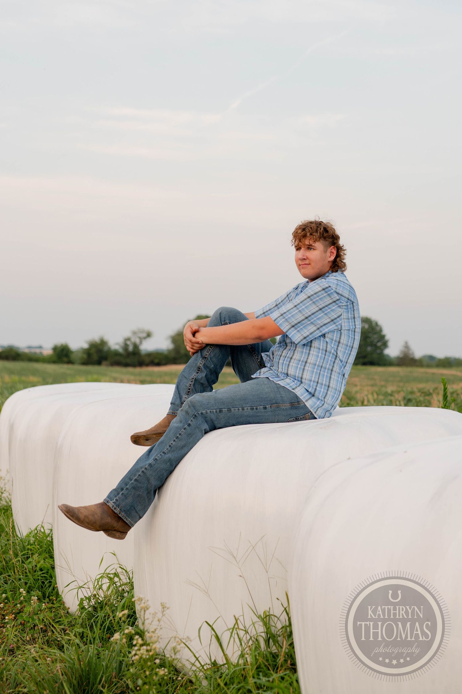 A person reclines on a white hay bale in a grassy field at dusk.