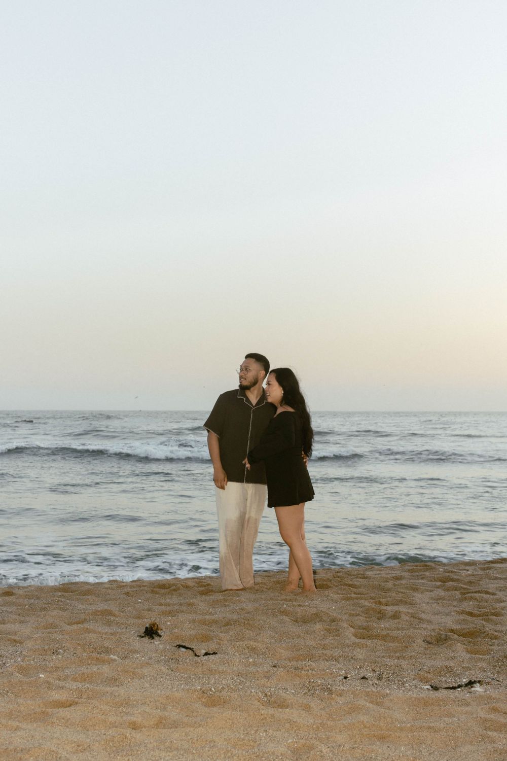 A romantic couple stands together on a beach at sunset with ocean waves in the background.