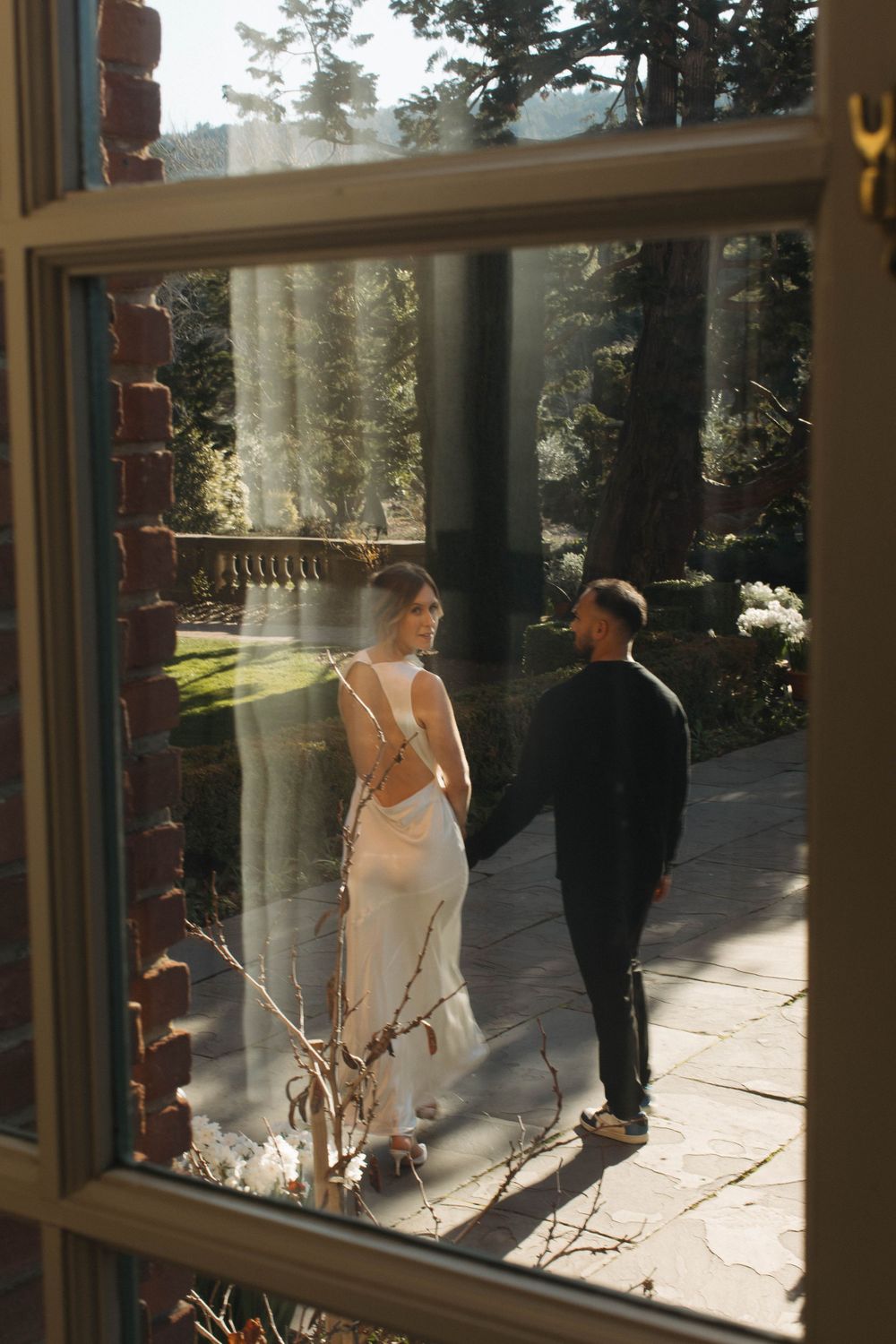 Couple walking hand in hand along a stone path at golden hour, viewed through a window frame.