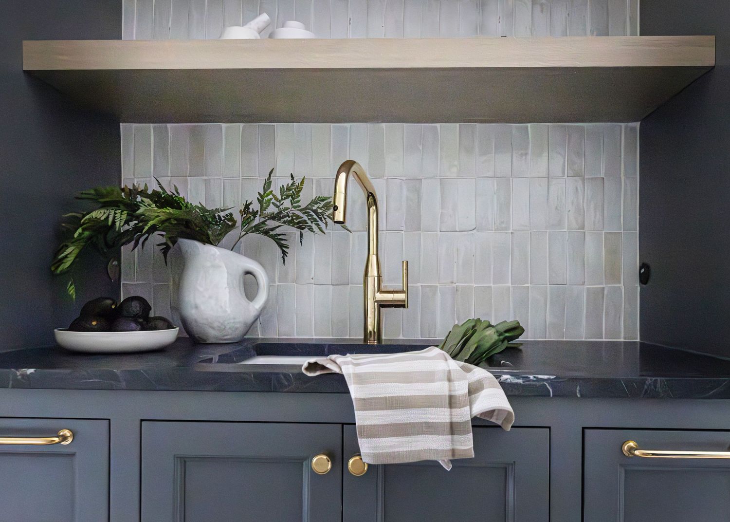 Modern kitchen with gray cabinets, brass fixtures, white ceramic tile backsplash, and potted plant on dark countertop.