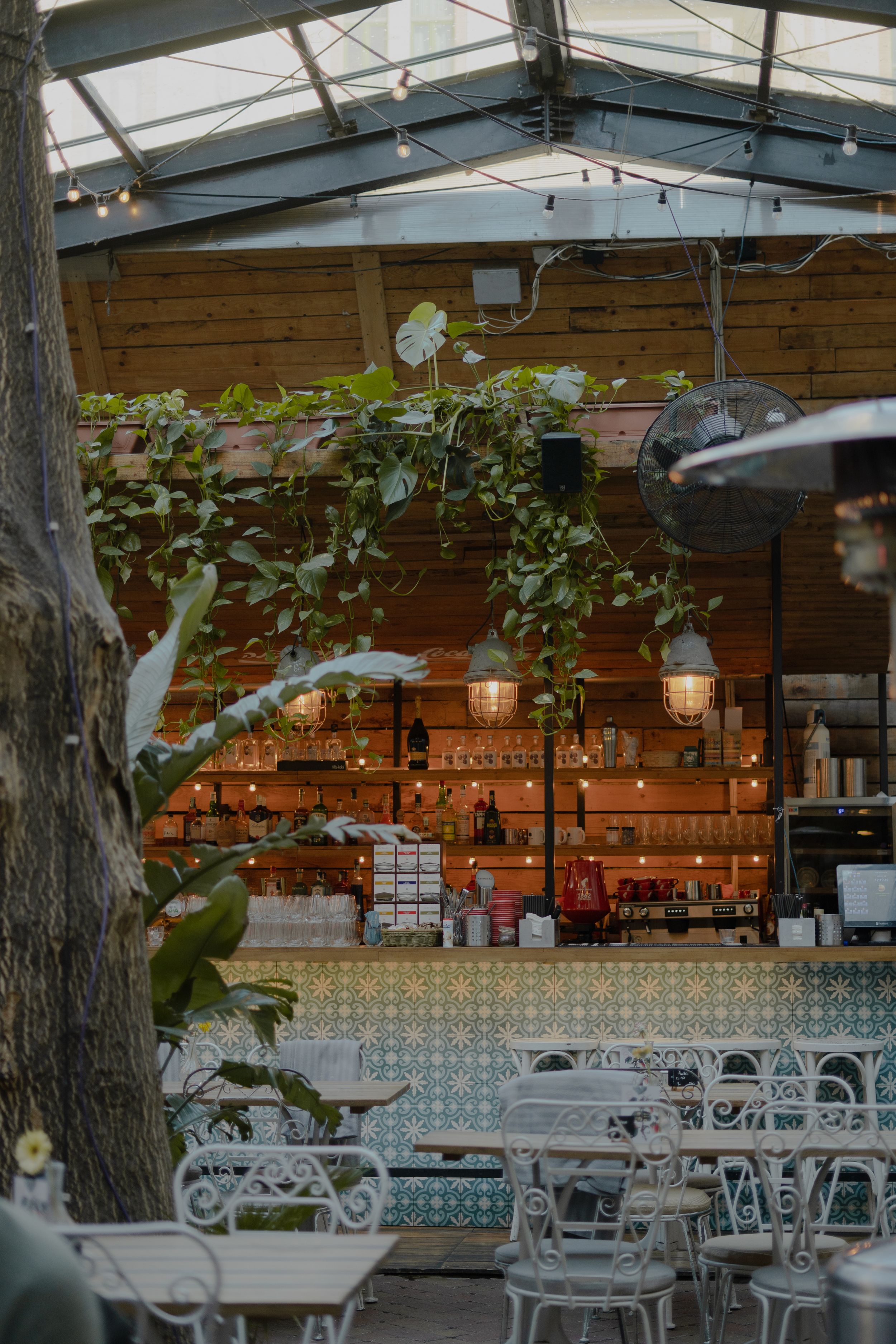 Cozy outdoor restaurant patio with white wrought iron furniture, string lights and greenery under a covered pergola.