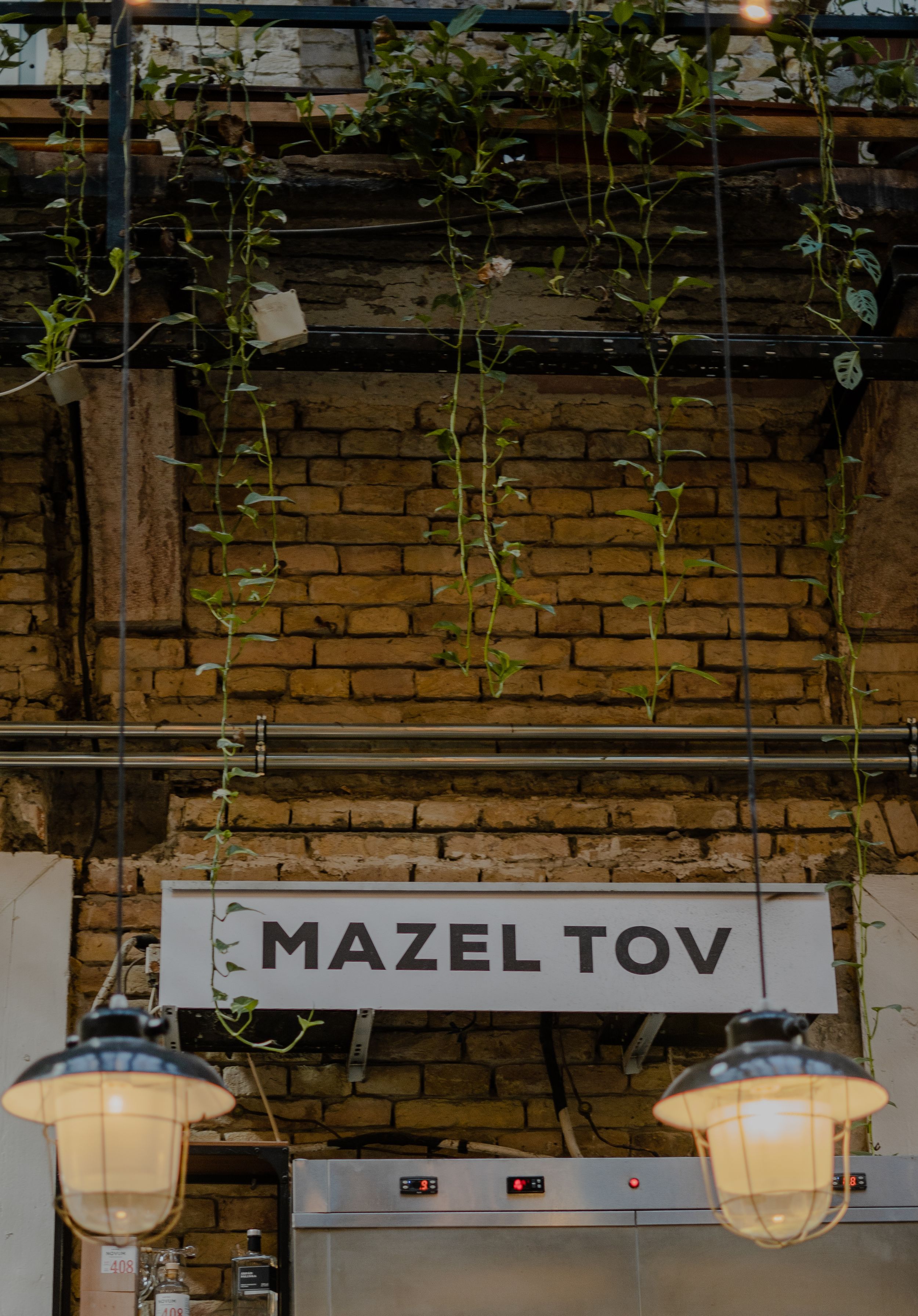 A rustic brick wall features a 'Mazel Tov' sign with hanging lanterns and climbing vines in a warm, inviting setting.