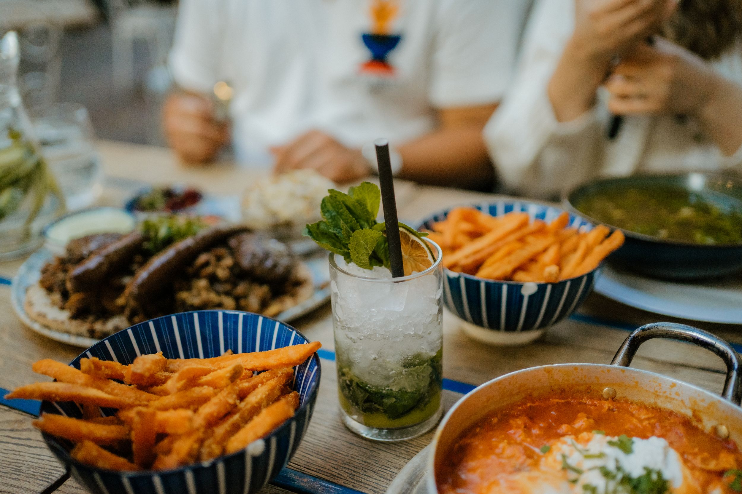 A restaurant table spread with french fries in blue striped bowls, mojito cocktail, and creamy orange soup garnished with herbs.
