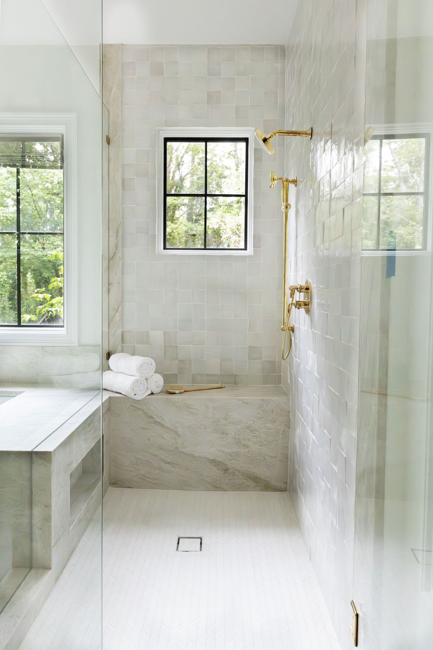 Modern marble bathroom with brass fixtures featuring a glass shower enclosure, white tiles and bright windows overlooking greenery.