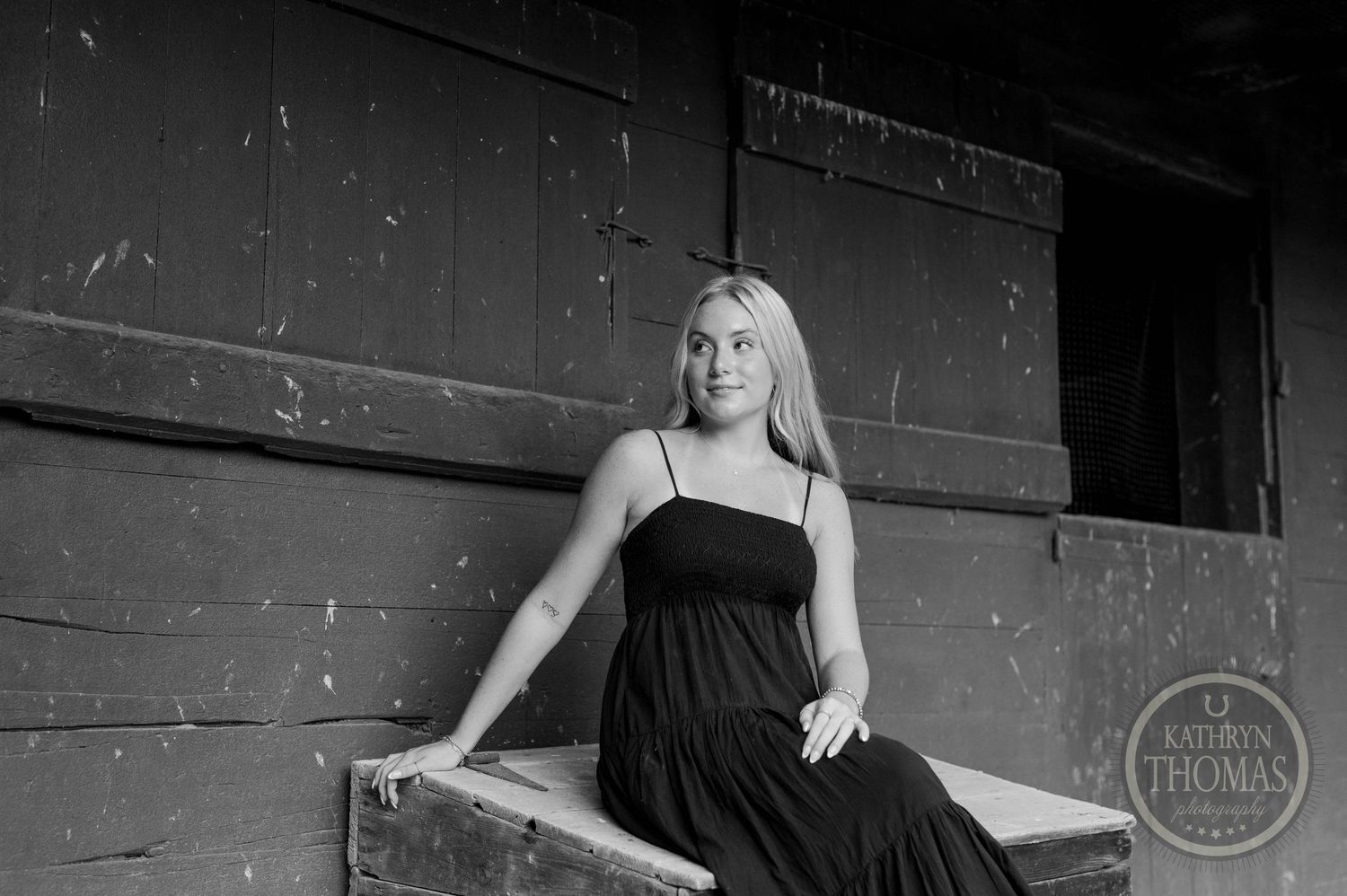 Black and white portrait of someone seated on a ledge wearing a dark dress in a rustic setting.
