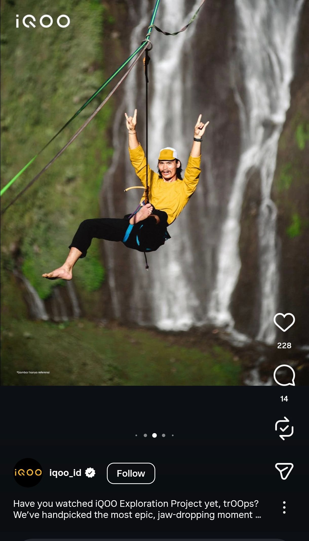Person in yellow jacket swinging on rope near a tall waterfall in lush green landscape.