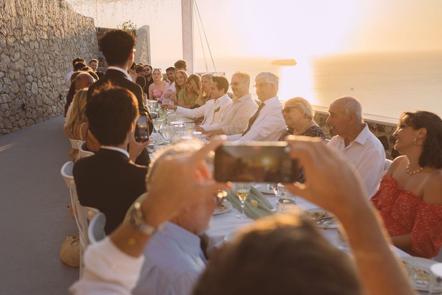 A destination wedding party enjoys an outdoor wedding speech at sunset while someone captures the moment on their phone.