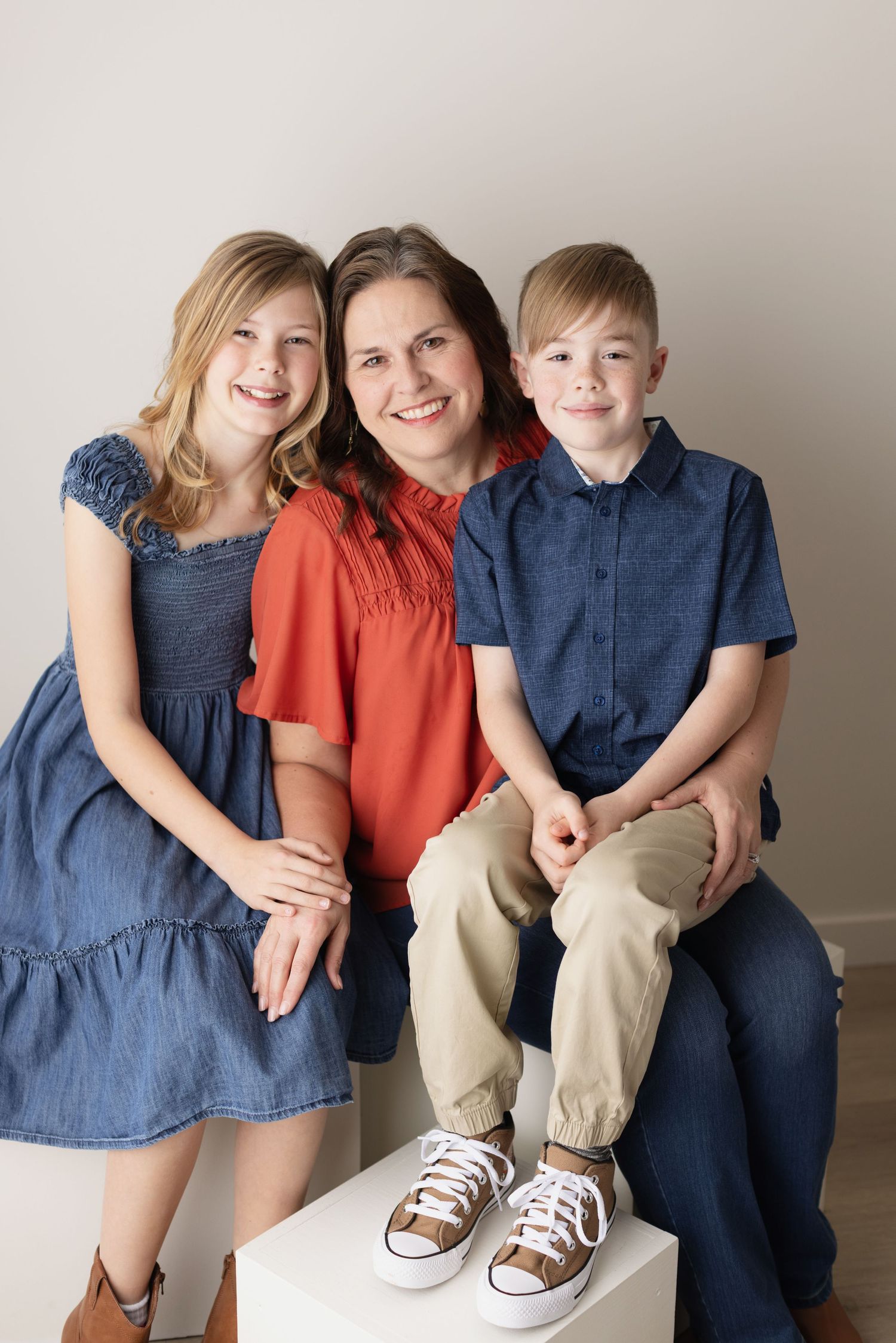 Family portrait in coordinating blue and orange outfits sitting together on a white pedestal against neutral background. Elizabeth Lang Phot