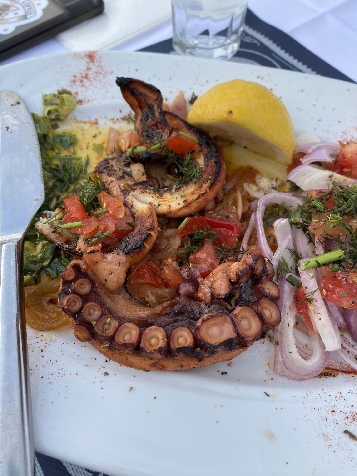 Grilled octopus tentacles served with lemon and fresh herbs on a white plate with red spice garnish.