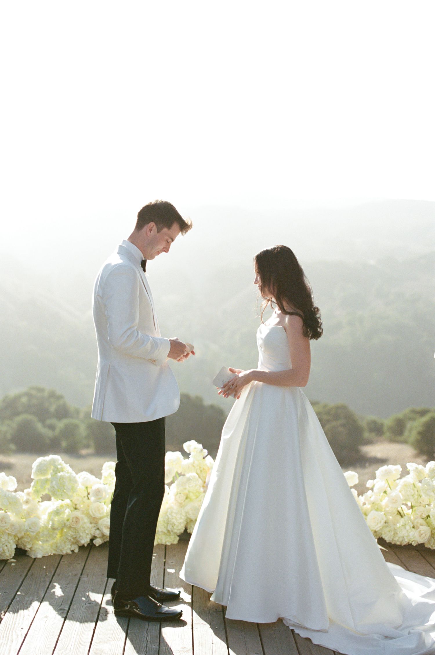 A couple in wedding attire dance together on an outdoor deck with white flowers and misty mountains in the background.
