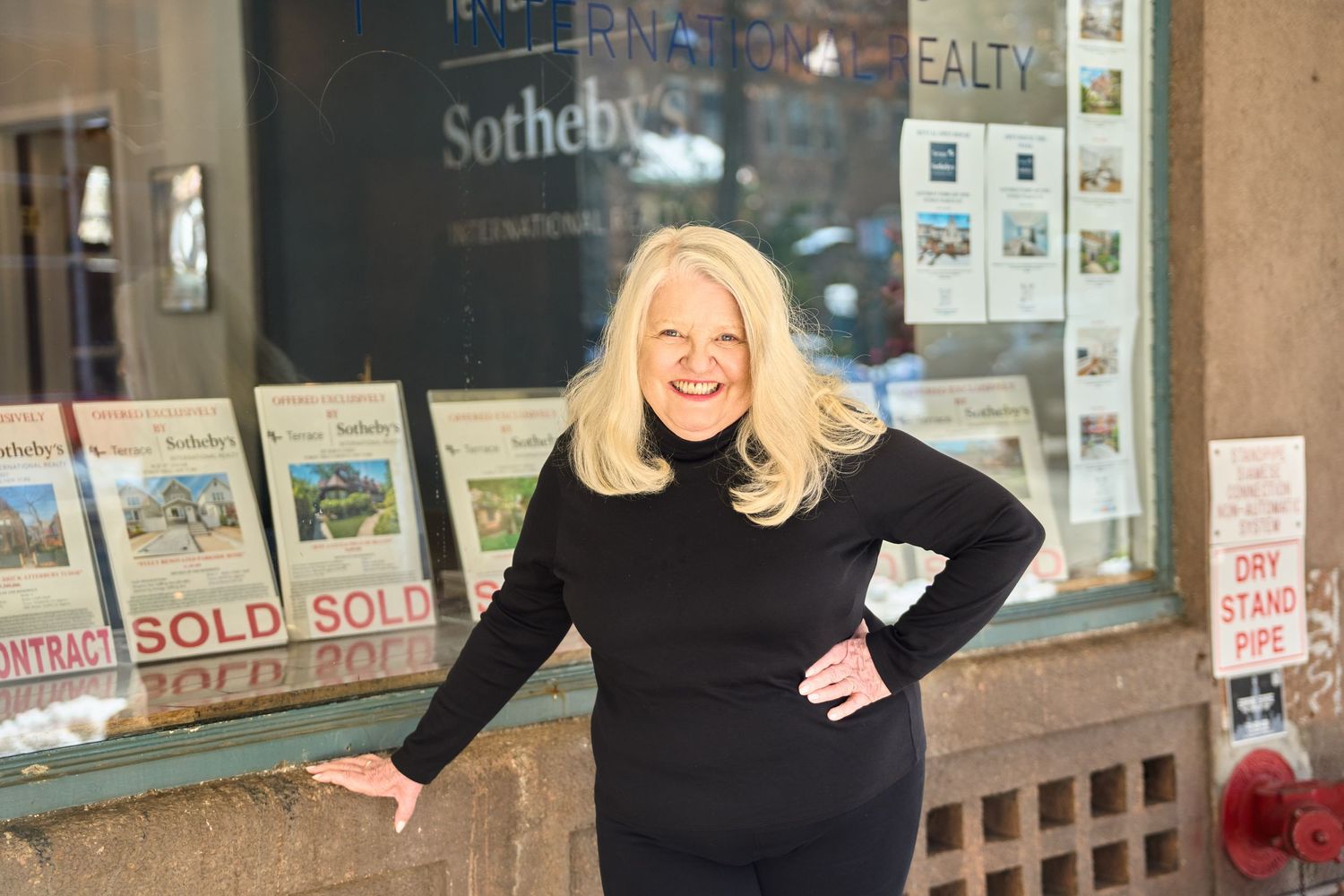 A real estate broker in a black sweater stands outside a storefront with glass windows and business signage.