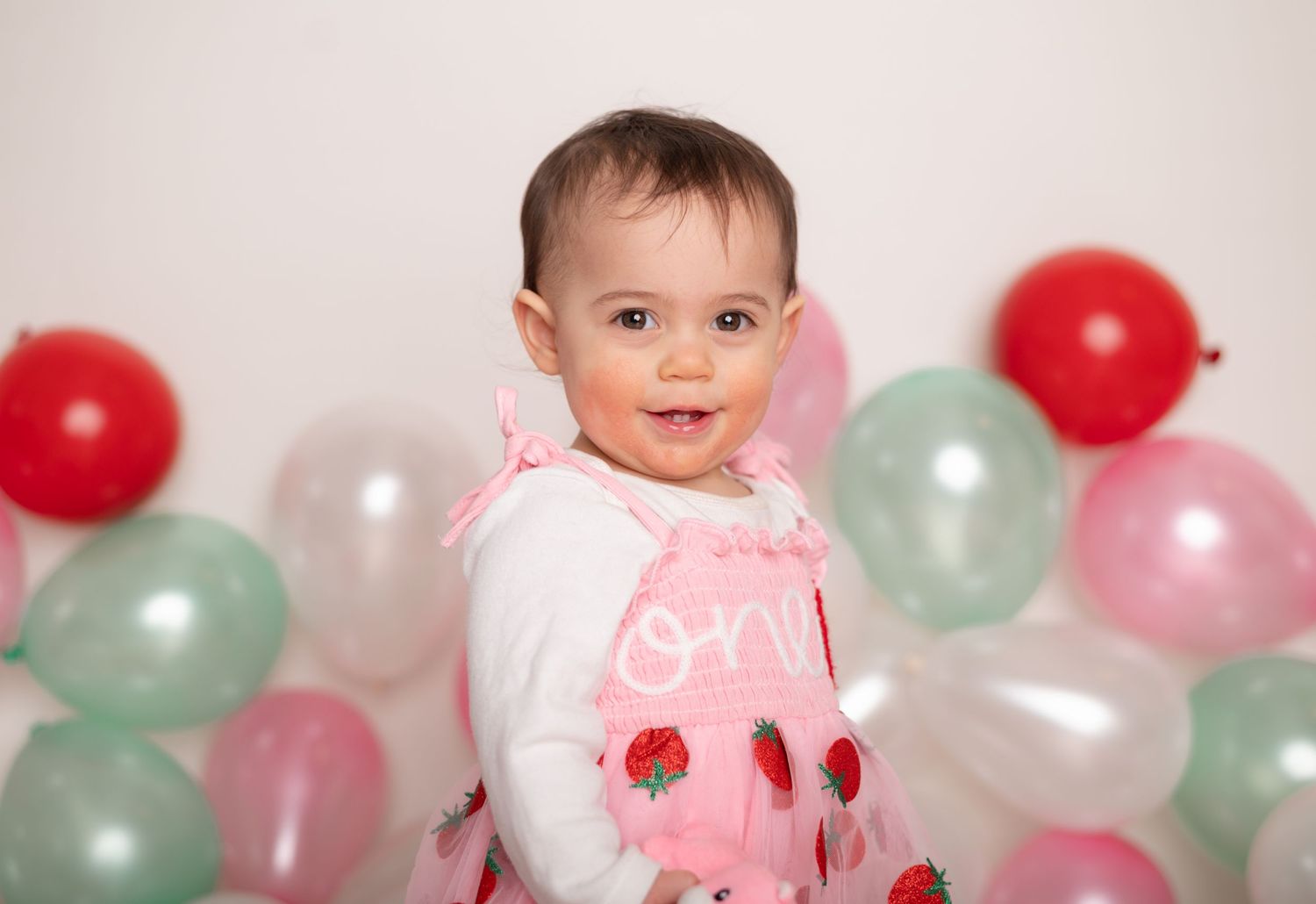 A baby in a pink and white outfit smiles while sitting among red, white, and mint green balloons.