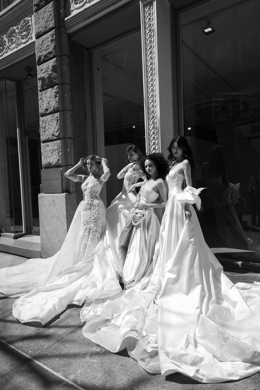 A group of people in elegant white wedding gowns with long trains pose together outside a building.