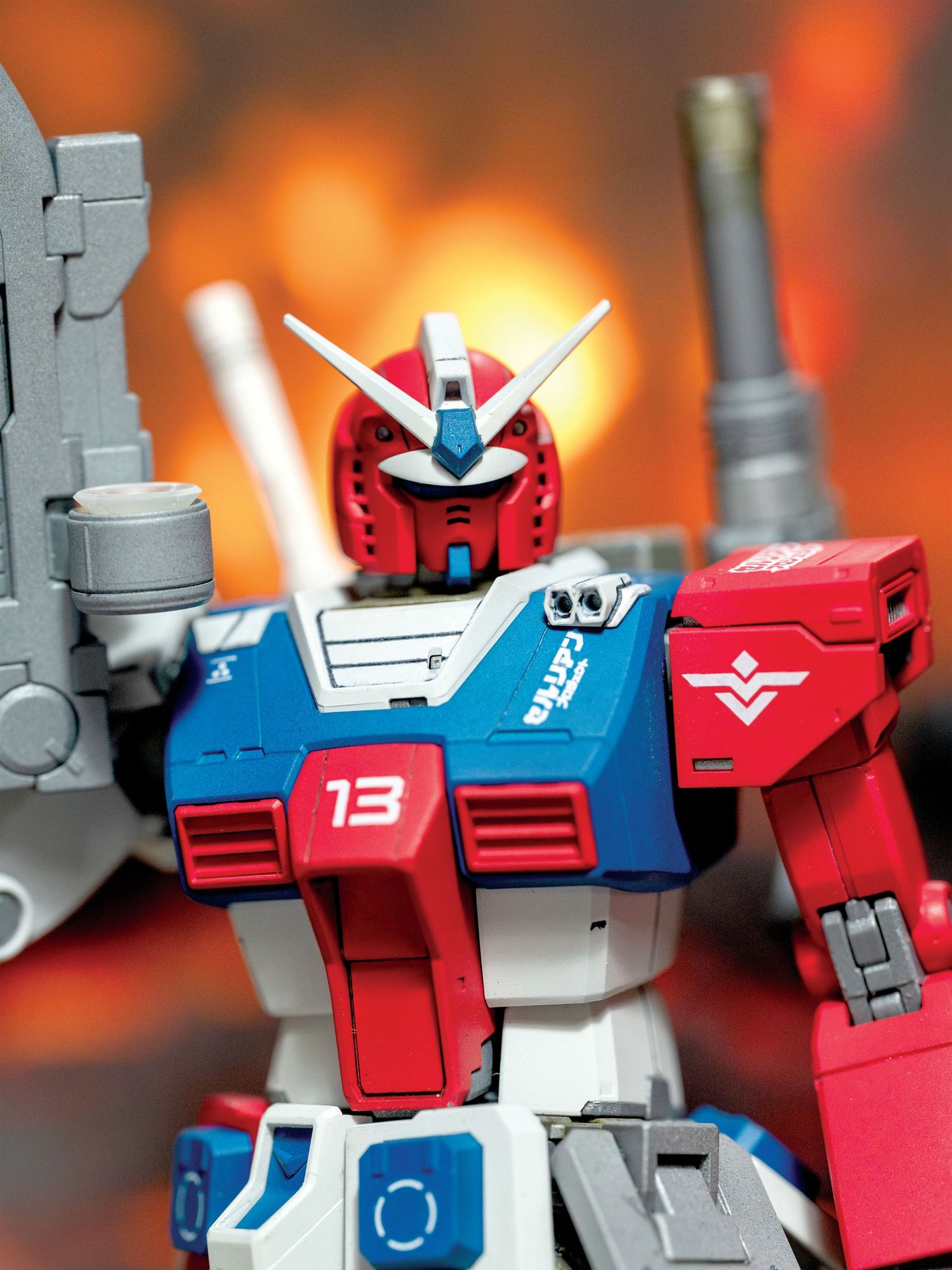A red and blue Gundam model figure shown in multiple poses against an orange blurred background.
