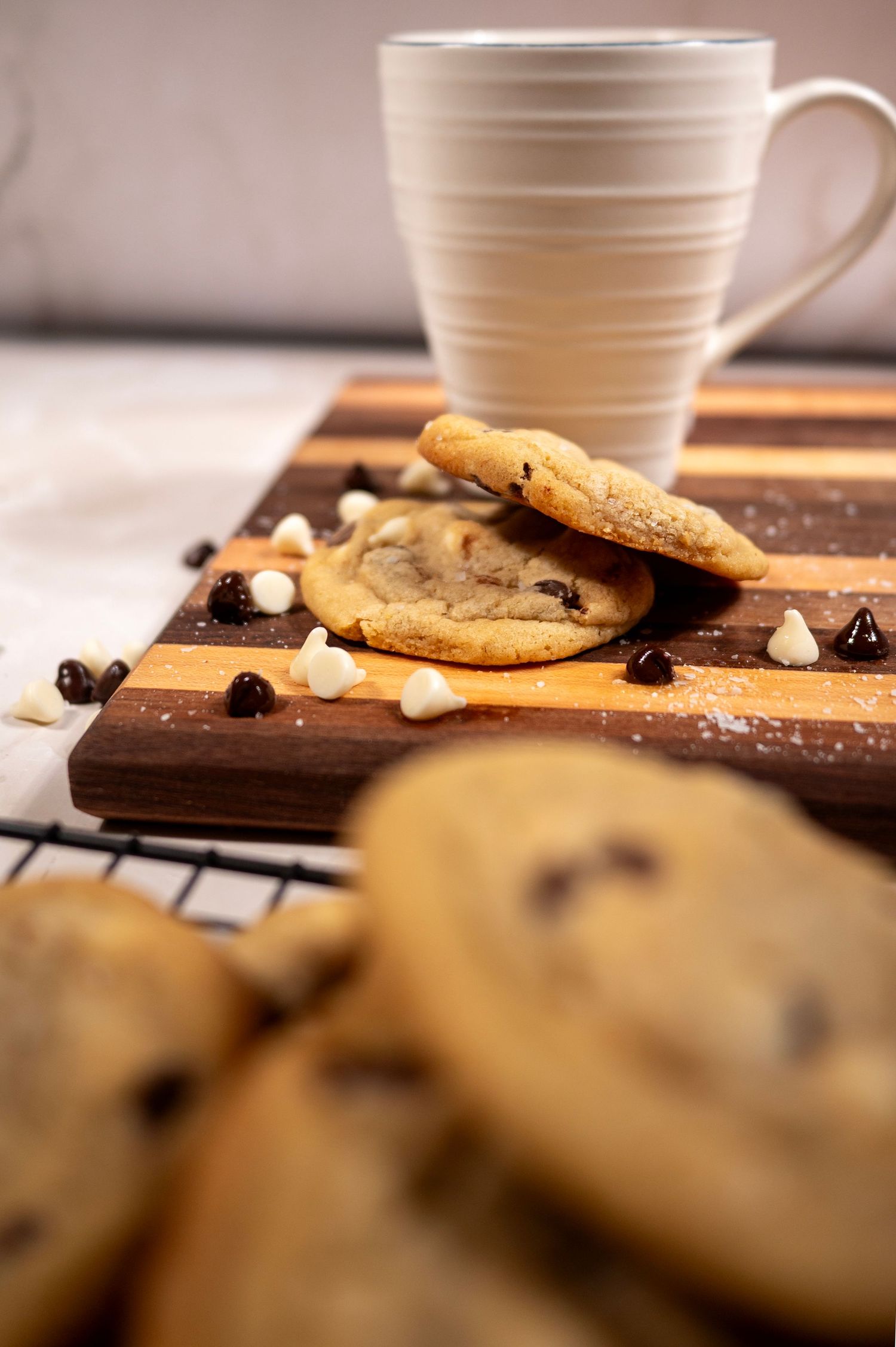 Chocolate chip cookies with white chocolate chips sit beside a white coffee mug on a rustic wooden surface.