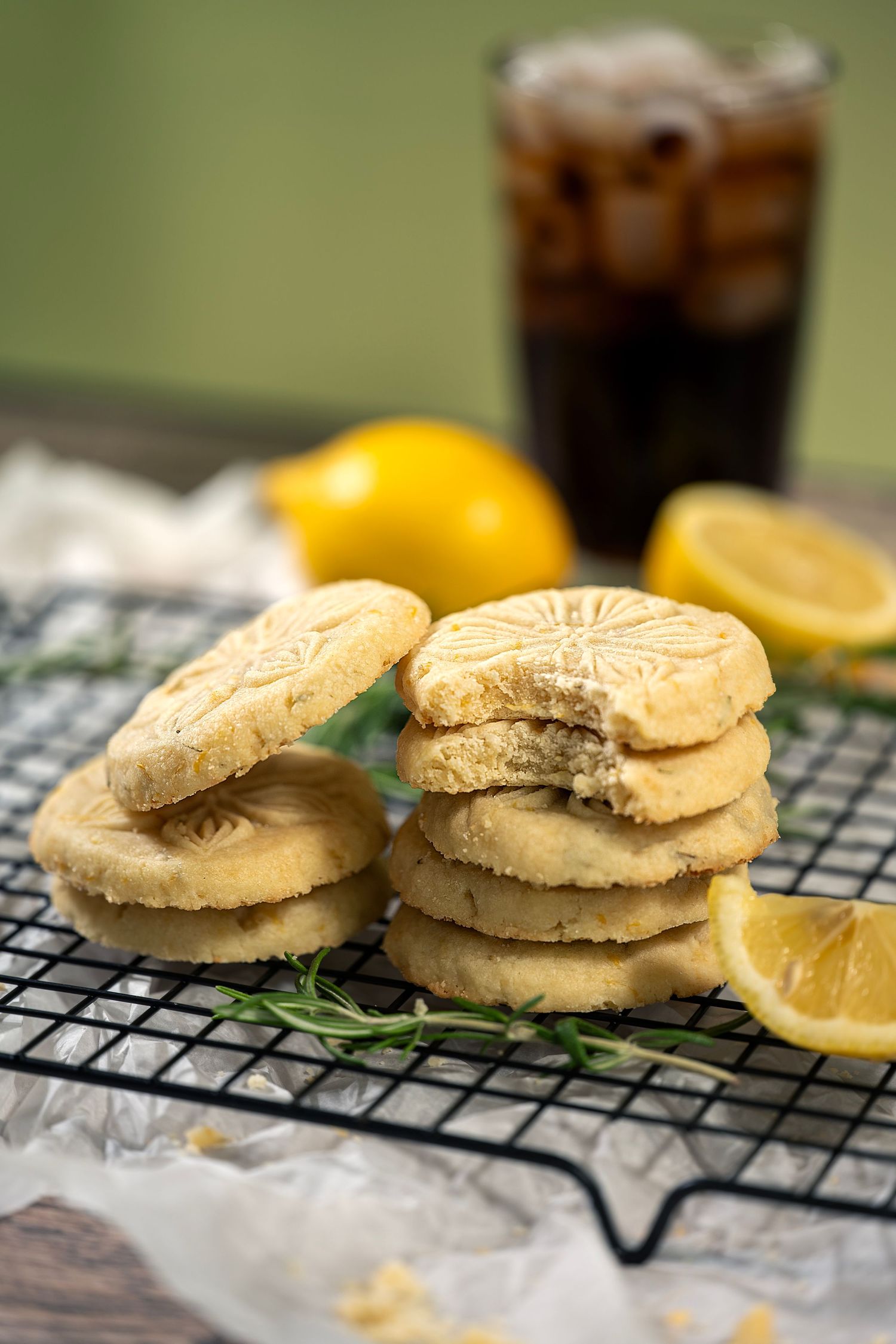 Freshly baked lemon shortbread cookies stacked in pairs on a cooling rack with citrus garnish and dark beverage.