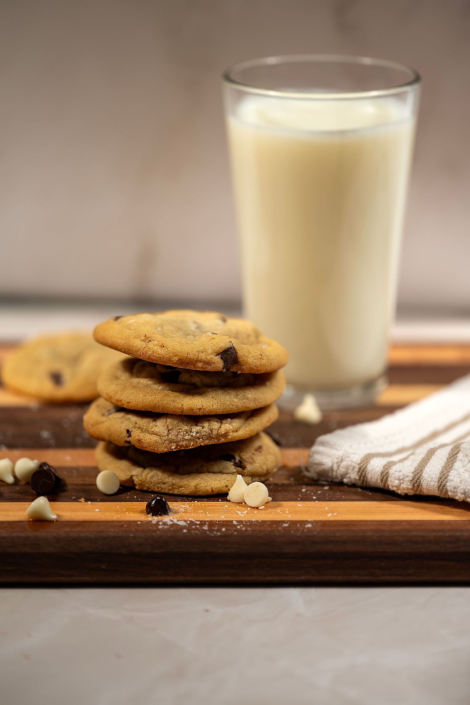 Stacked chocolate chip cookies with crumbs on a wooden board next to glasses of milk in warm lighting.