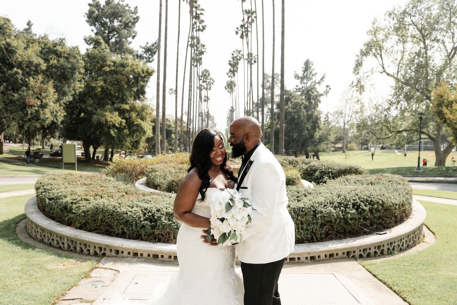 A couple shares an intimate moment in a lush garden setting surrounded by white pillars and greenery.