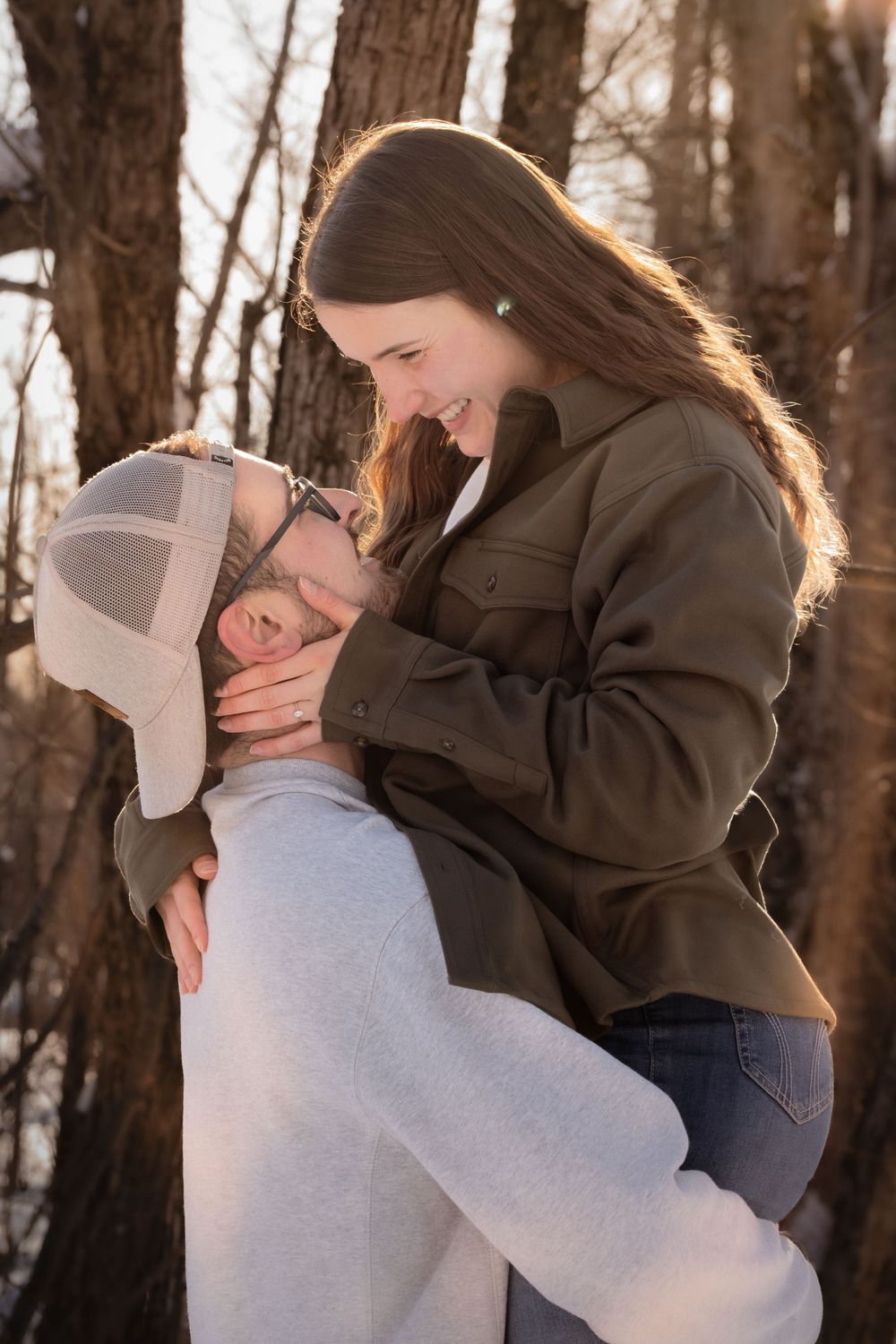 A romantic outdoor winter portrait shows a couple embracing and laughing together against a backdrop of bare trees.