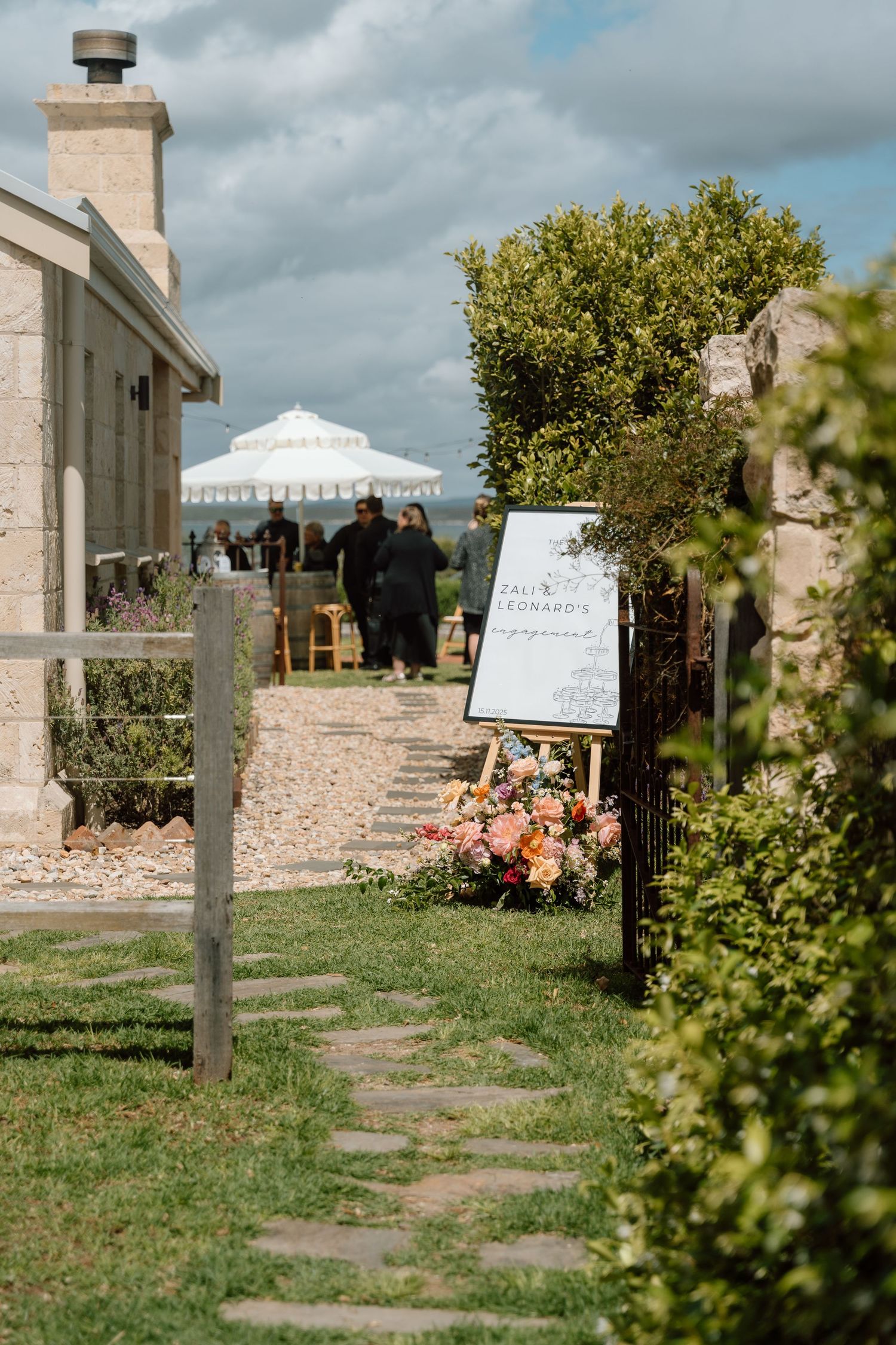 Stone entrance gate with decorative pillars leads to a garden path under dramatic cloudy skies.