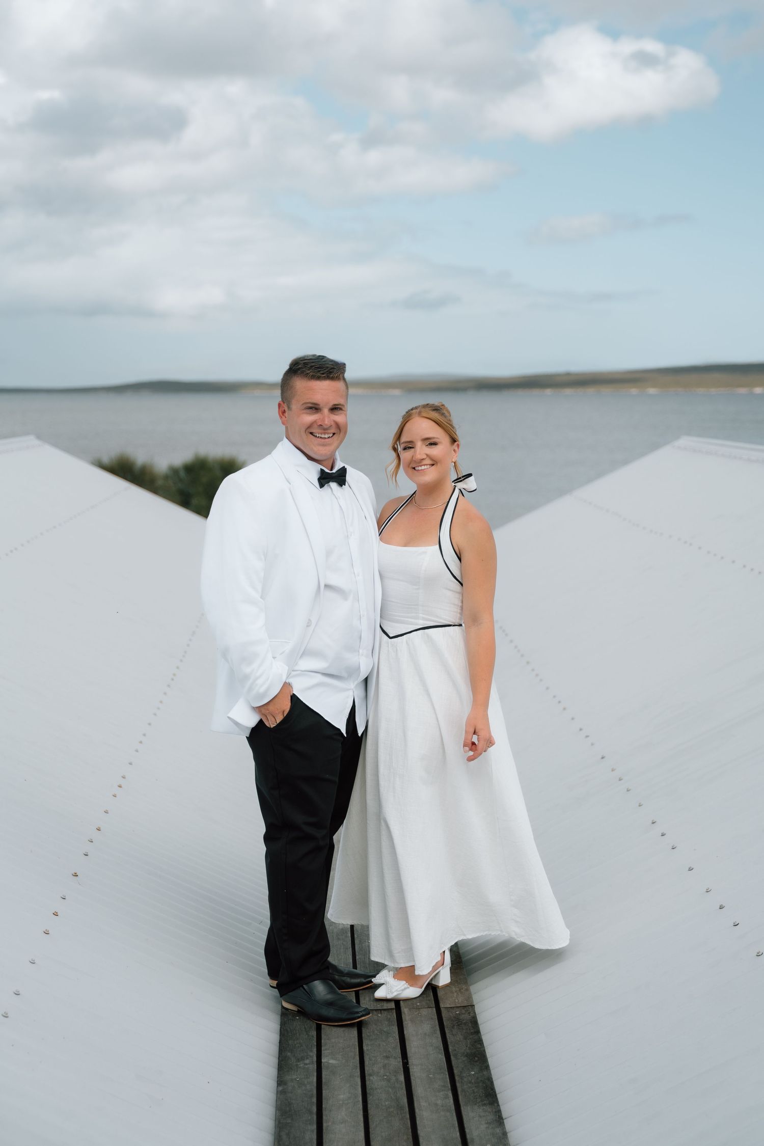 A couple in formal wedding attire stands on a white rooftop deck overlooking a coastal waterway.