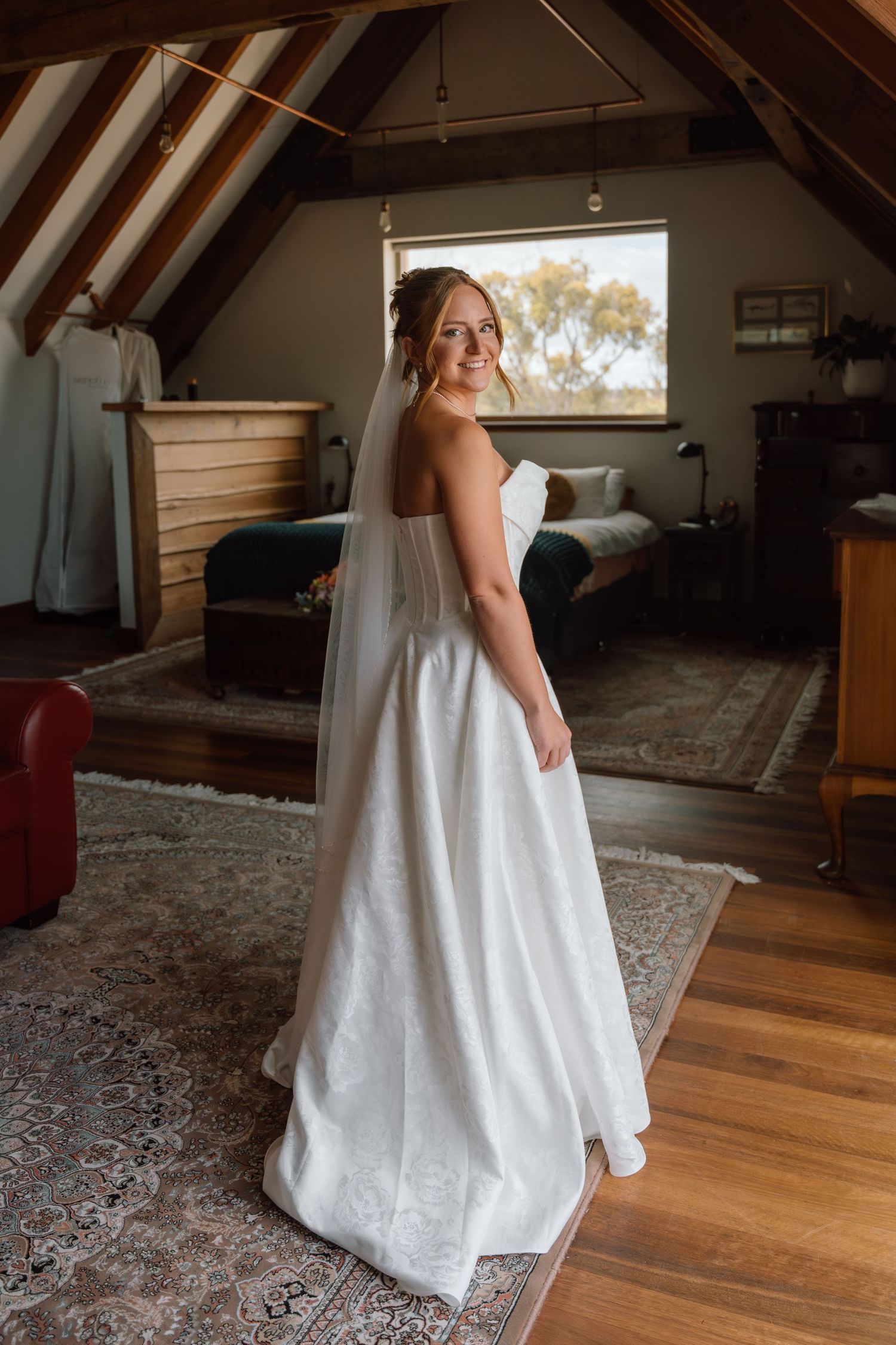 A bride in a flowing white wedding dress stands in a rustic attic bedroom with wooden beams and natural light.