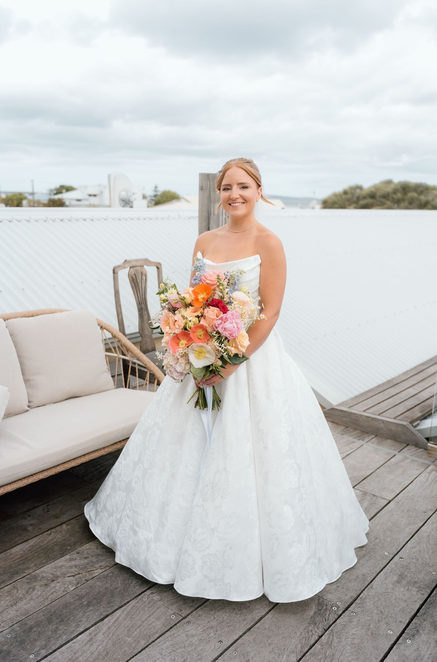 A bride in a classic white ballgown holds a vibrant coral and peach bouquet on a rooftop deck.