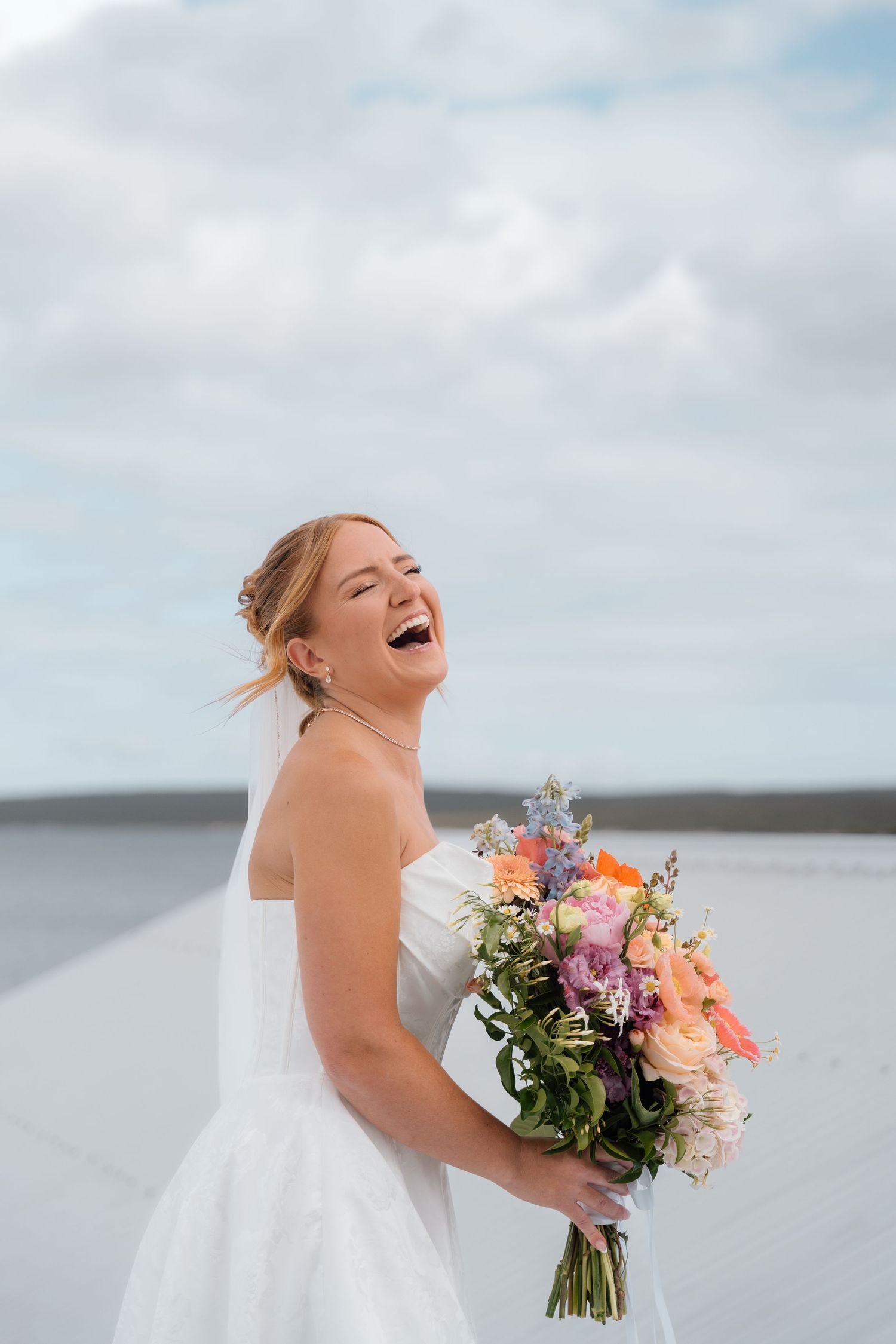 A joyful bride in a white dress laughs while holding a vibrant bouquet against a cloudy sky backdrop.