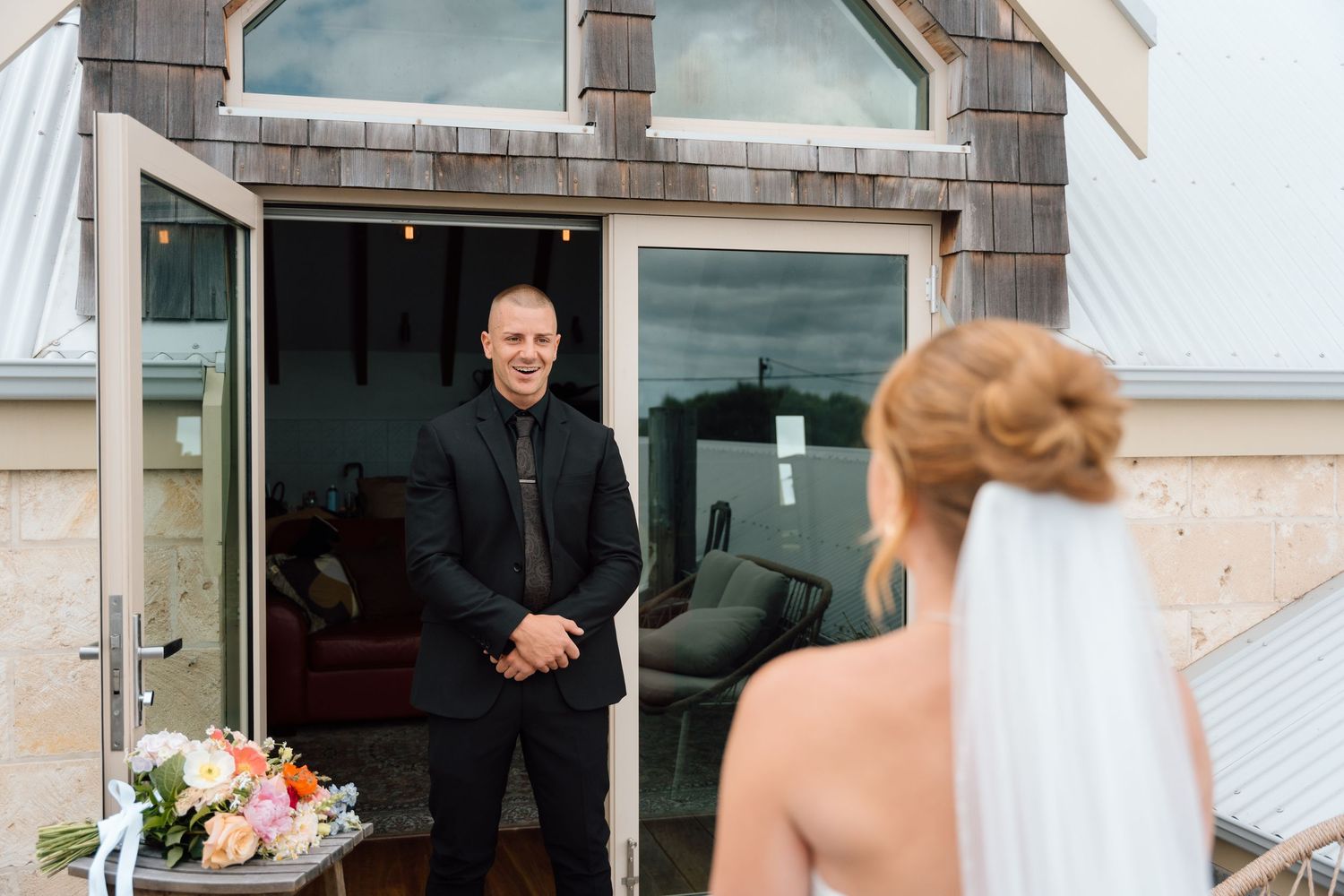 A wedding party gathers outside a brick building with shingled roof on a sunny day.