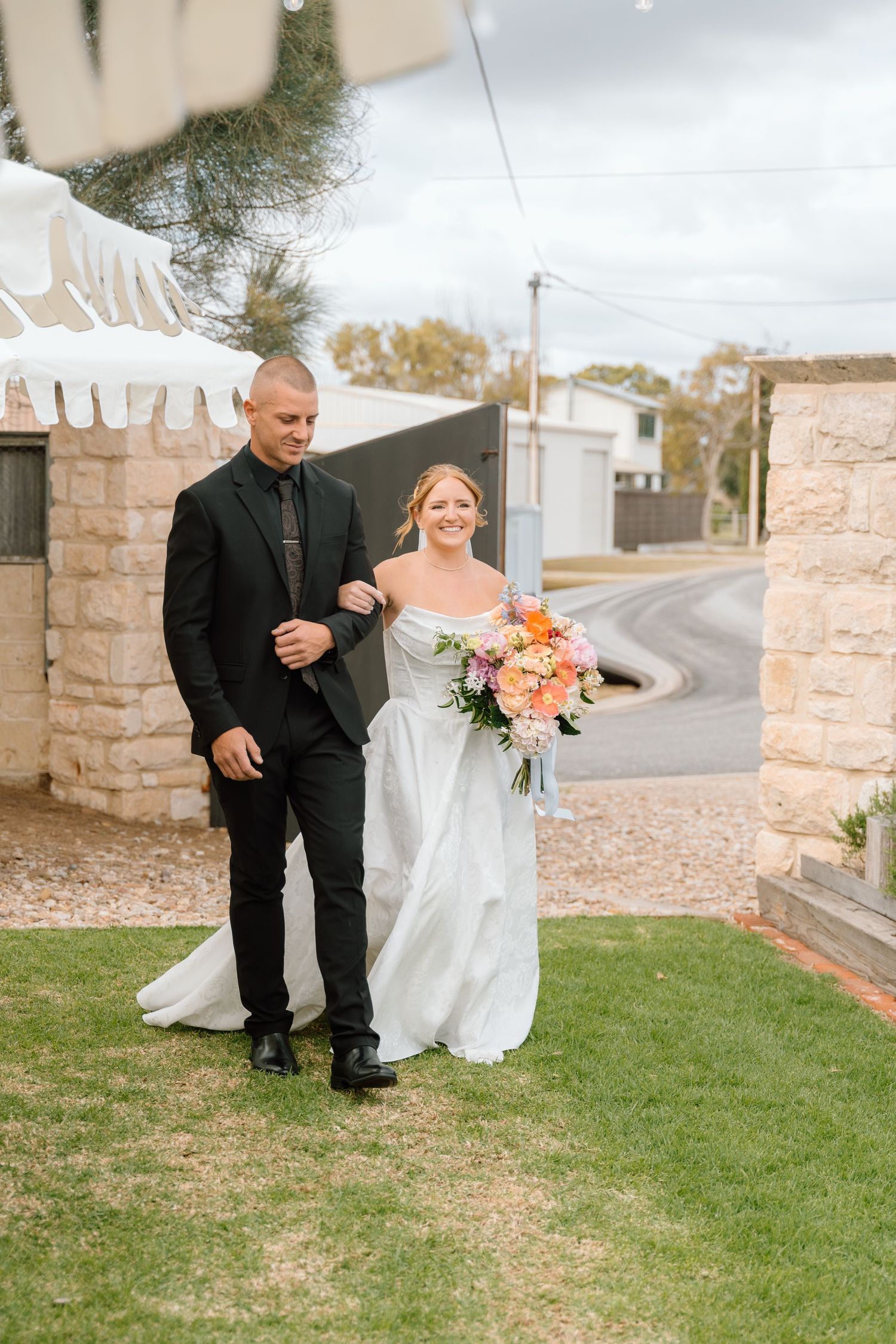 A wedding ceremony on a grassy lawn with stone walls, featuring a couple walking arm in arm with coral bouquet.