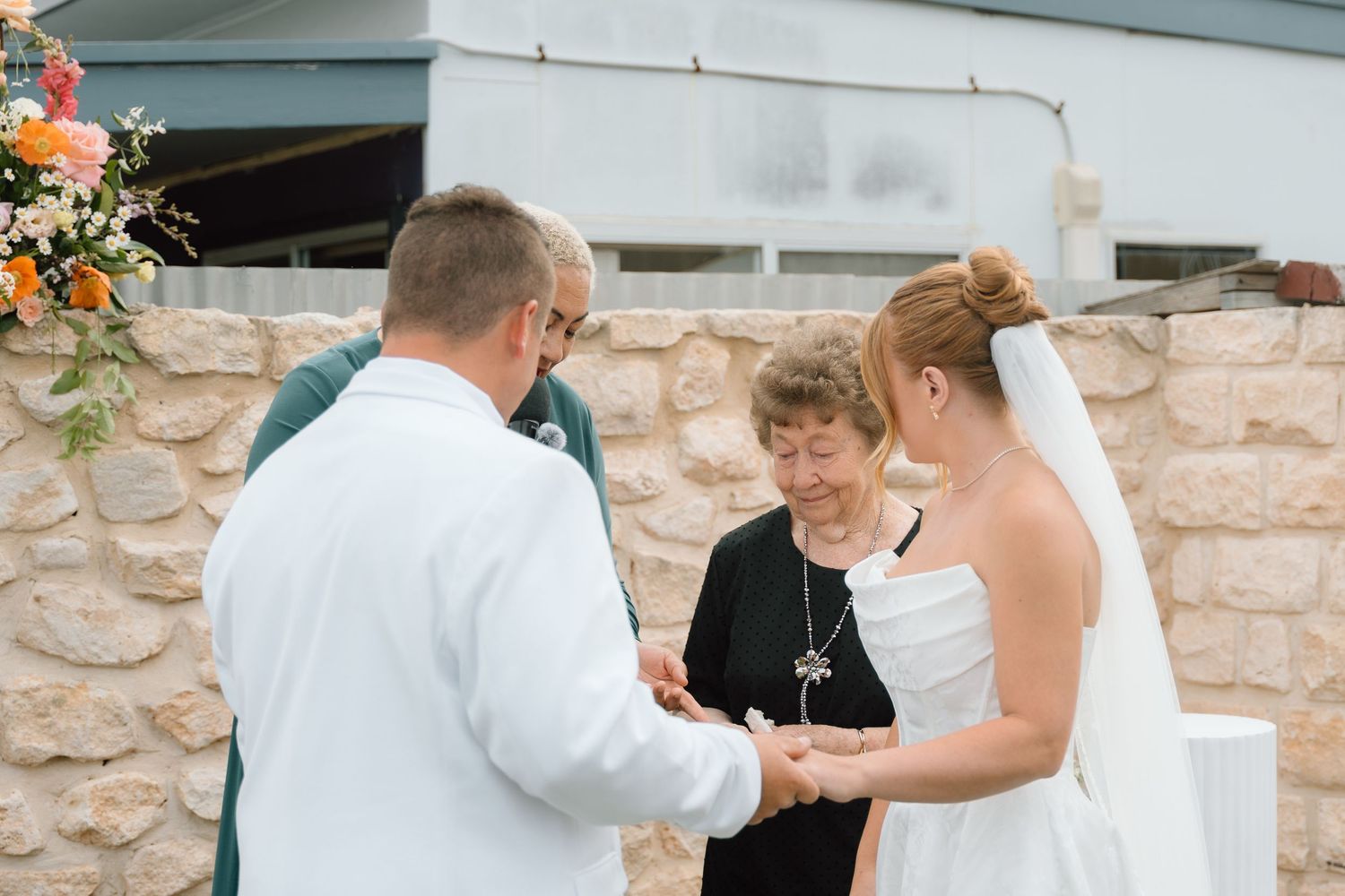 A wedding ceremony takes place outdoors with an officiant and couple exchanging vows near a stone wall with flowers.
