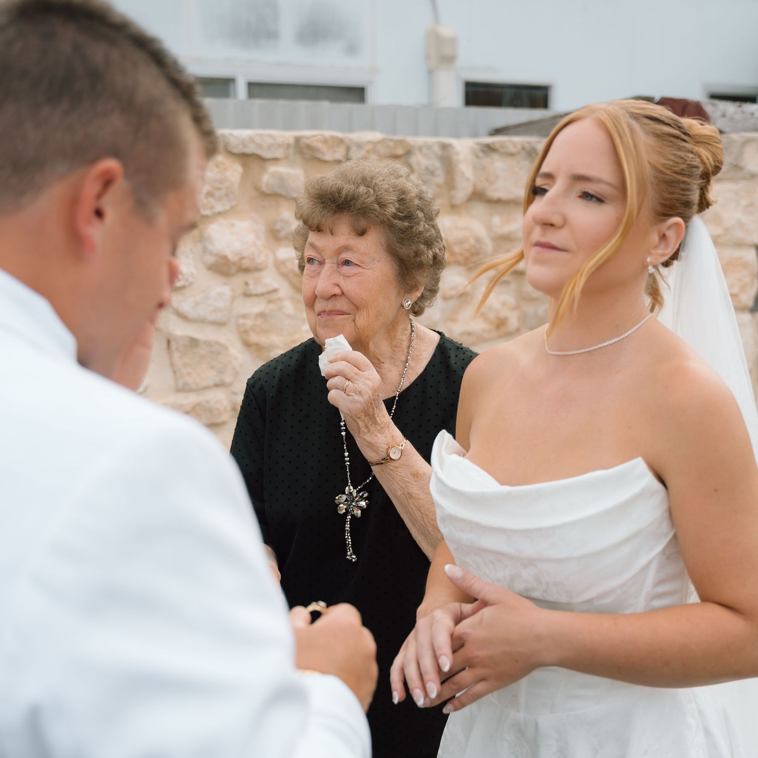 A wedding ceremony moment captured with participants in white attire and an elderly guest in black.