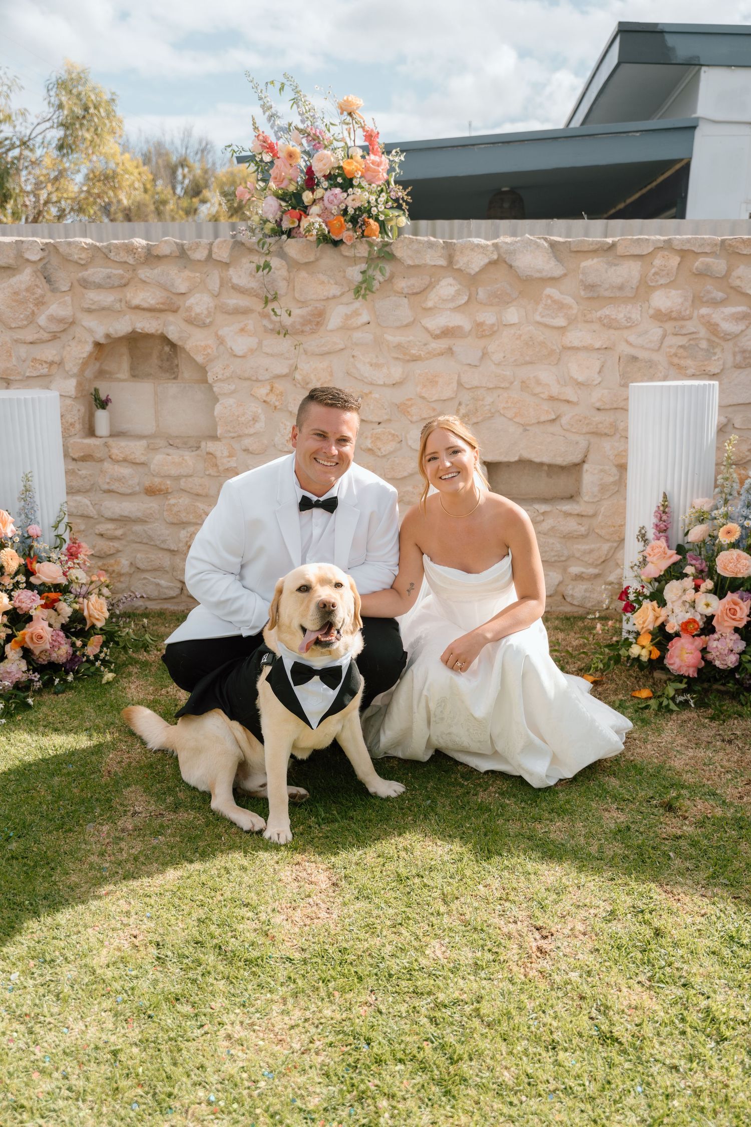 A couple in wedding attire sits on grass with their dog wearing a bowtie near pink floral decorations and stone wall.