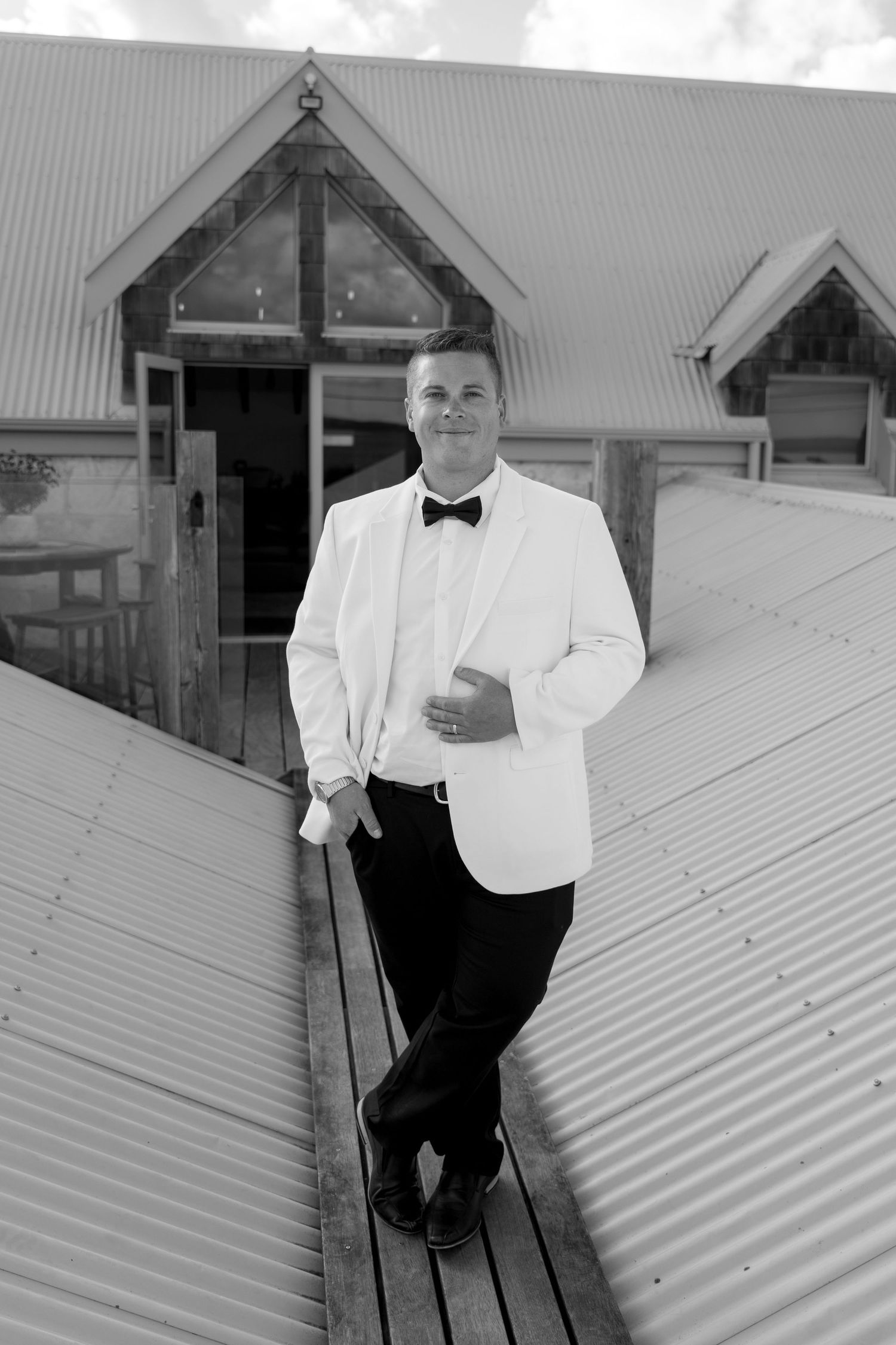 A person in a white dinner jacket and bow tie stands on a corrugated metal roof near a peaked architectural structure.