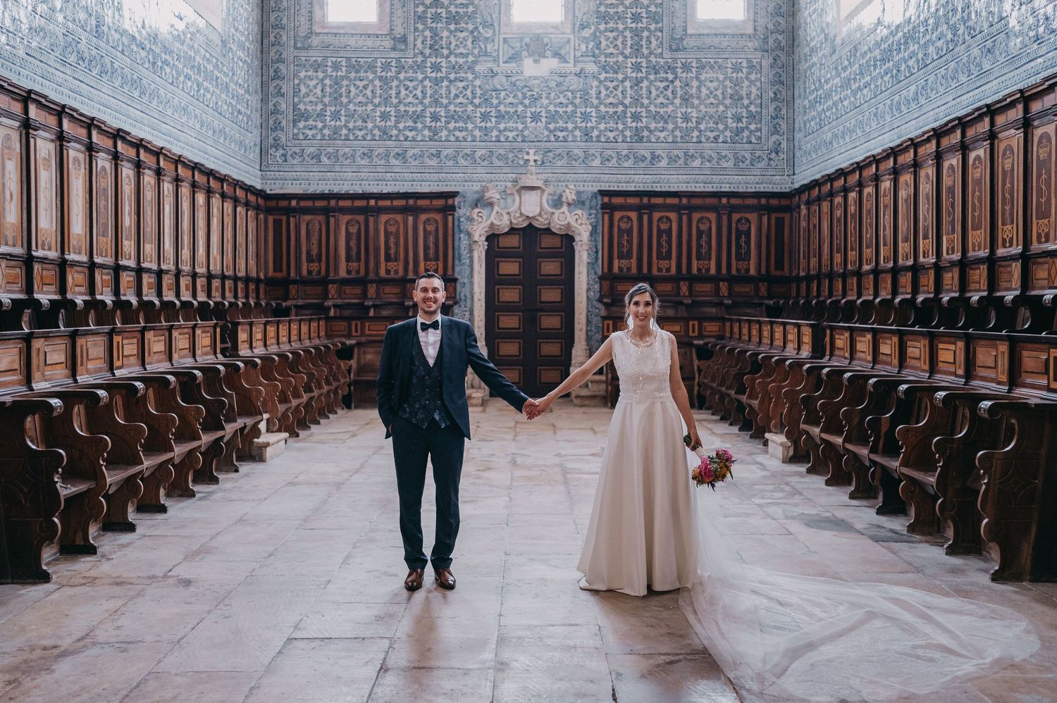 A couple walks down a long church aisle lined with wooden pews and ornate architectural details.