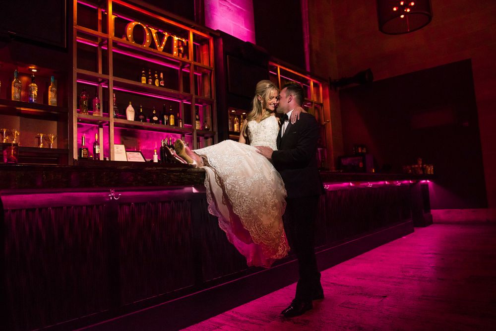 Newlyweds share their first dance in a ballroom illuminated by dramatic pink and purple lighting.