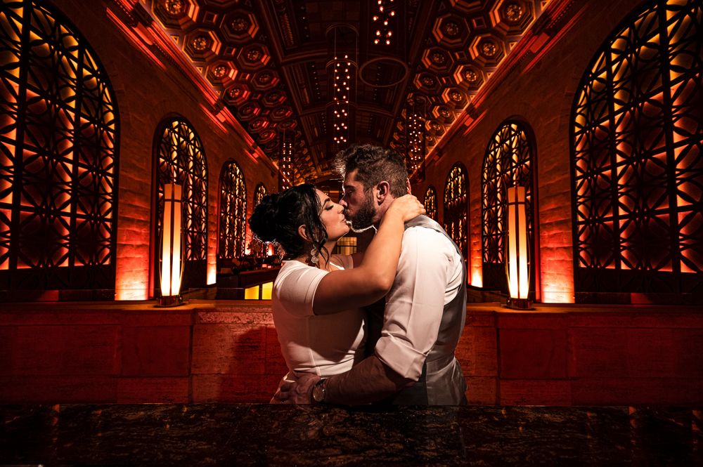 A romantic couple embraces in a dramatic red-lit venue with ornate architectural details and glowing lights.